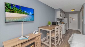 A compact hotel room with a TV displaying a beach scene, a small kitchenette, a wooden dining table, and a bed. - Sea Mist 51408 (Myrtle Beach)