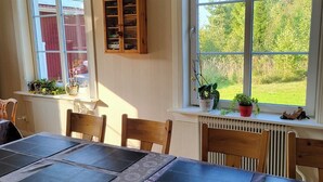 A dining area with a table set for six, wooden chairs, a window with a view of greenery, and a wooden cabinet above the window.
