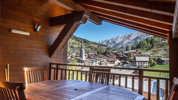 Una terraza de madera con una mesa y sillas, con vistas a un pueblo montañoso.