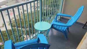 Un balcon avec des chaises bleues et une petite table, donnant sur une plage parsemée de parasols et où se trouvent des gens.