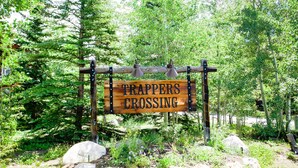 Puinen kyltti, jossa lukee ”TRAPPERS CROSSING”, sijoitettu luonnonmukaiseen ympäristöön, jossa on puita ja kiviä.