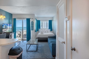 A room with a bed, a sofa, a table, a trash bin, and a view of the sea through a sliding door.