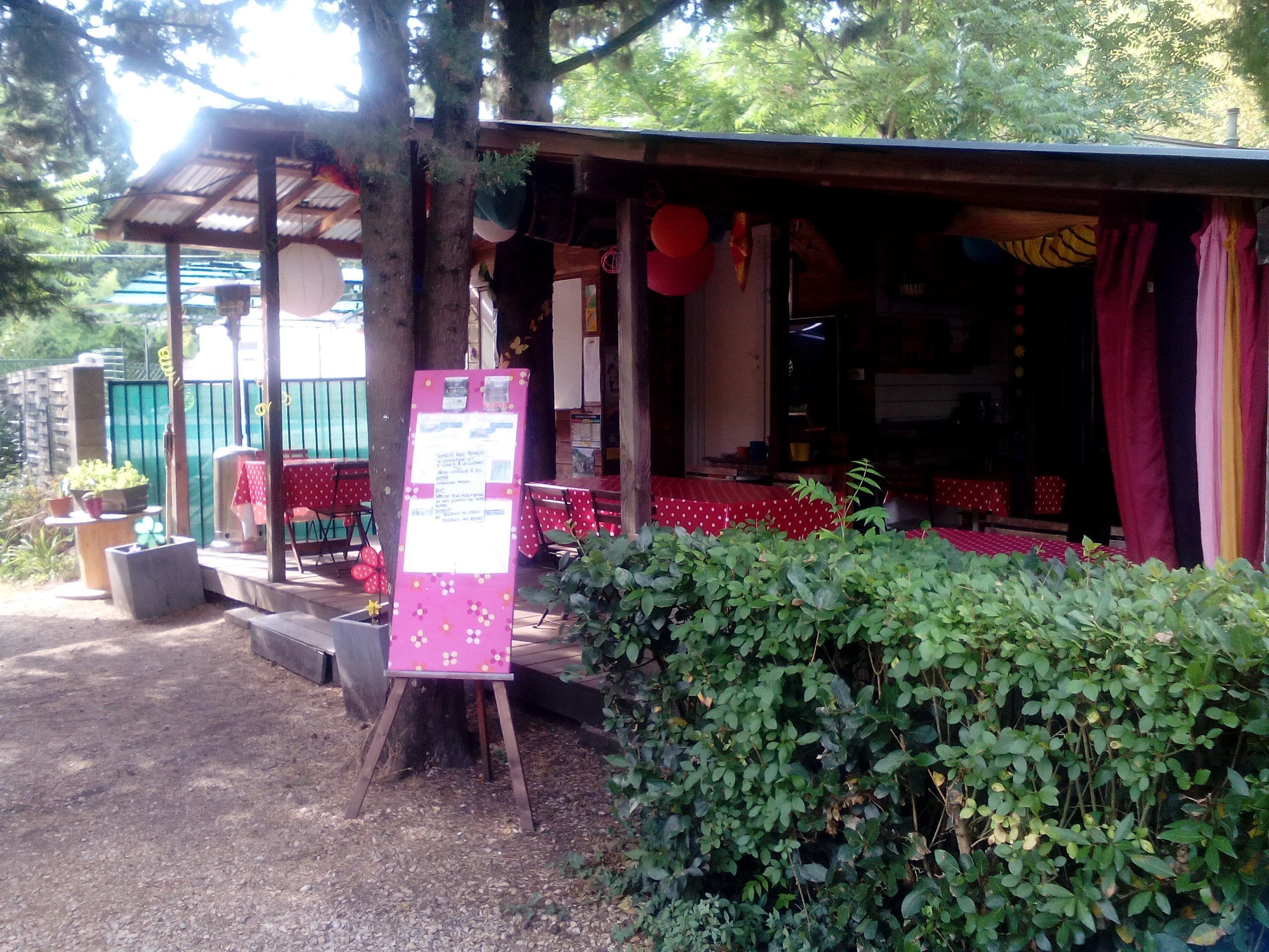 Ein rustikaler Außenbereich mit Sitzgelegenheit, einer Holz-Pergola, rot-weißen Karotischdecken und einem Schwarzebrett mit Aushängen.