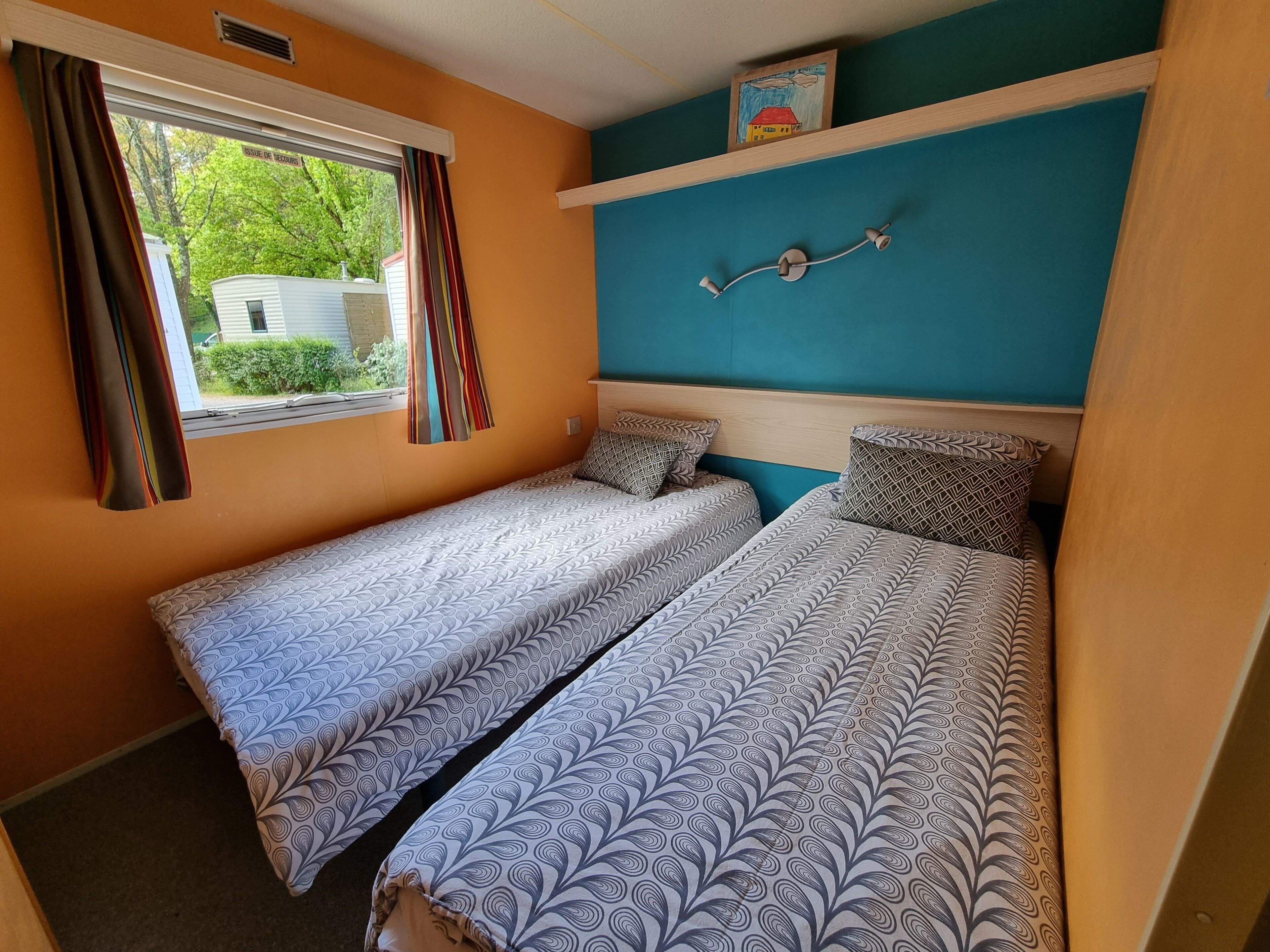 Une chambre avec deux lits, une fenêtre avec des rideaux, un luminaire fixé au mur et un petit tableau au-dessus du lit.
