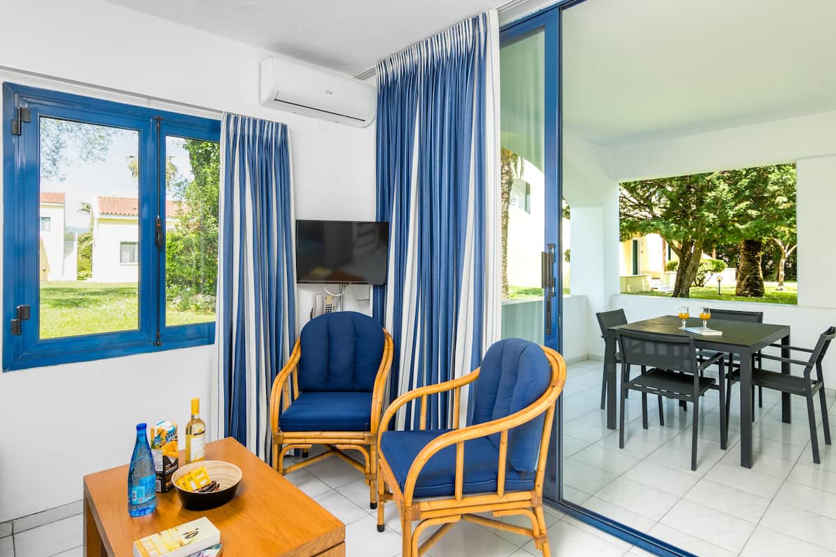 Ein Zimmer mit Fernseher, zwei blauen Stühlen, einem Esstisch mit Stühlen und Blick auf Grünfläche.