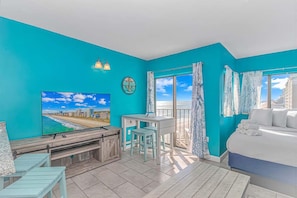 A room with teal walls, a bed, a desk, a chair, a TV, and a balcony with curtains.