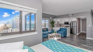 Een modern appartement met balkon, een woonkamer met bank, een keuken met roestvrijstalen apparatuur en een groot raam met uitzicht over de stad.