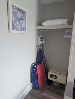 A small room with a bed, a framed picture on the wall, a suitcase, a ironing board, and a box with a remote control.