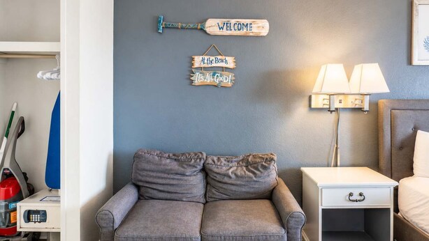 A room with a grey sofa, a nightstand with a lamp, and wall decorations including a 'WELCOME' sign and 'At the Beach' sign. - Sea Mist 50809 (Myrtle Beach)