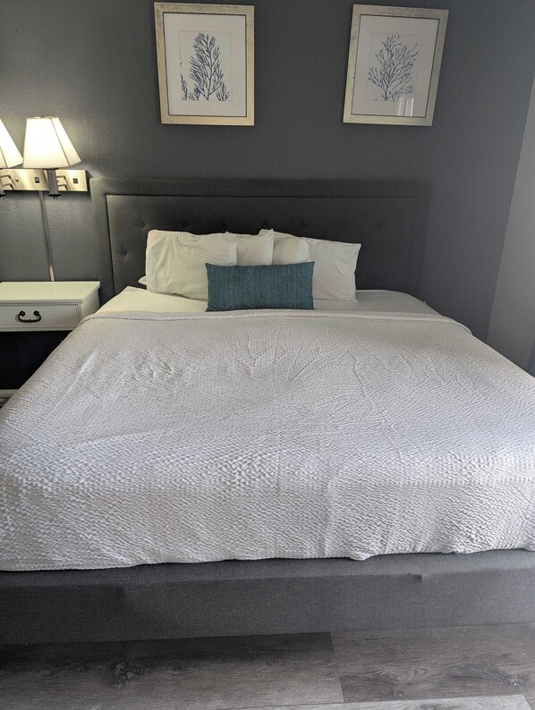 A neatly made bed with a textured white cover, a dark headboard, and two framed botanical prints on the wall.