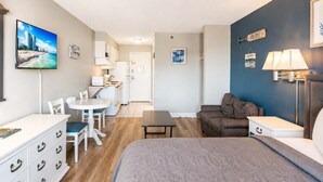 A compact hotel room with a bed, a sofa, a dining area with a table and chairs, a kitchenette, and a TV displaying a beach scene. - Sea Mist 50809 (Myrtle Beach)