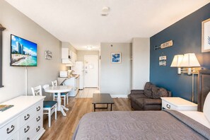 A compact hotel room with a bed, a sofa, a dining area with a table and chairs, a kitchenette, and a TV displaying a beach scene. - Sea Mist 50809 (Myrtle Beach)