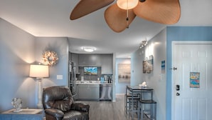 A modern living room with a leather chair, a dining table with two chairs, a kitchen with grey cabinets, and a ceiling fan. - Sea Pond Villa Unit 3 (North Myrtle Beach)