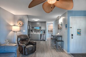 A modern living room with a leather chair, a dining table with two chairs, a kitchen with grey cabinets, and a ceiling fan. - Sea Pond Villa Unit 3 (North Myrtle Beach)
