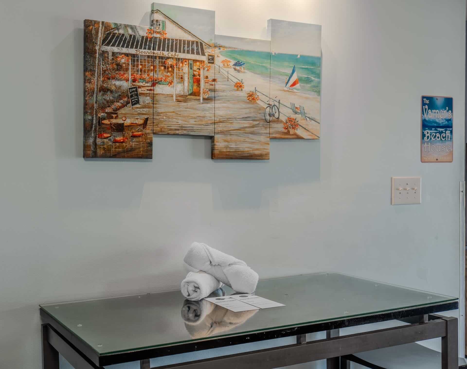 A wall with a beach-themed painting, a glass table with a rolled towel, and a light switch.