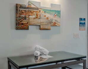 Un mur orné d’un tableau sur le thème de la plage, une table en verre avec une serviette roulée, et un interrupteur.