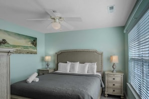 Une chambre à coucher avec un lit, deux tables de chevet avec des lampes, un ventilateur de plafond, un tableau représentant une scène de plage et une fenêtre avec des stores.