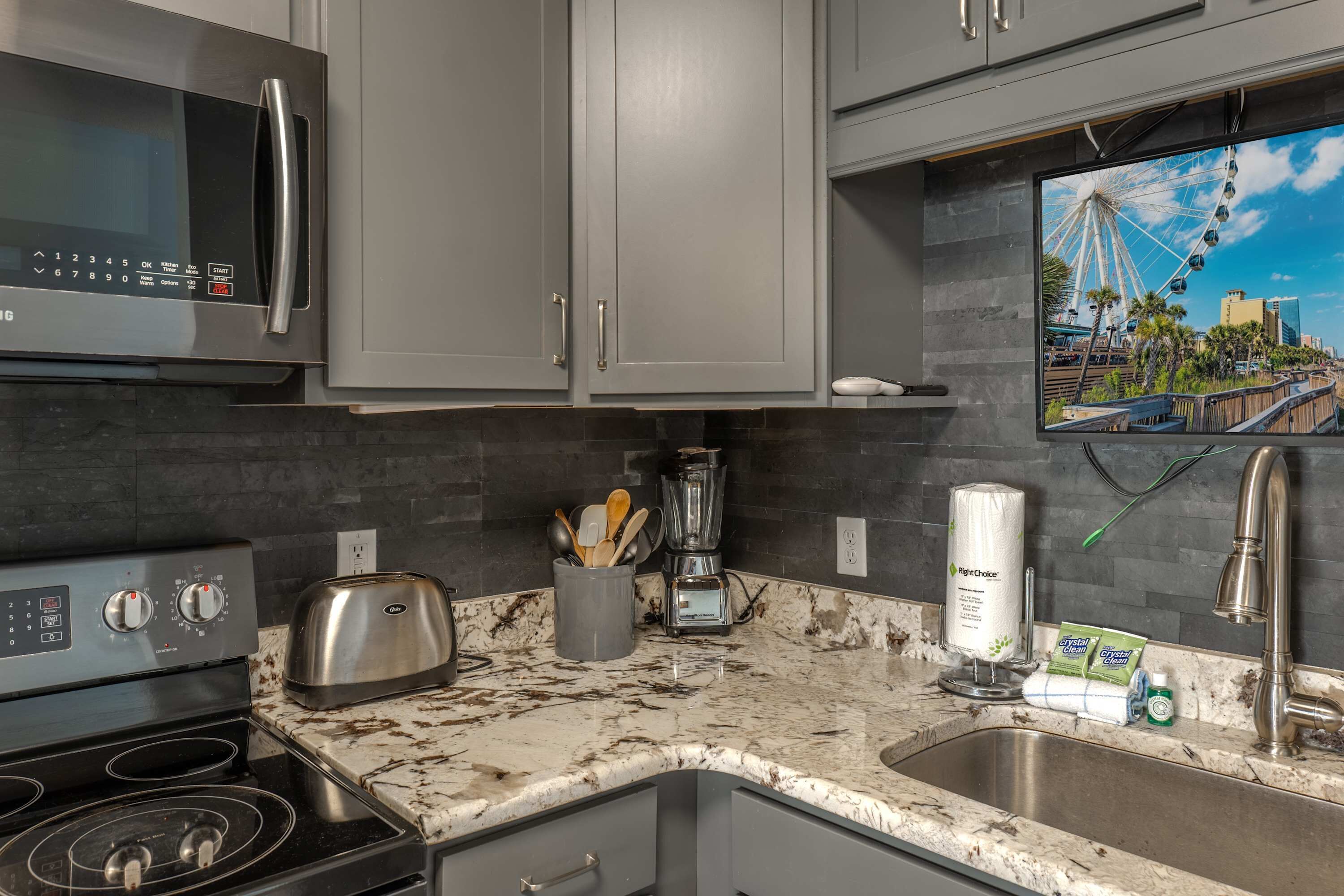 A kitchen with a microwave, toaster, stove, blender, and a large TV mounted above the counter.