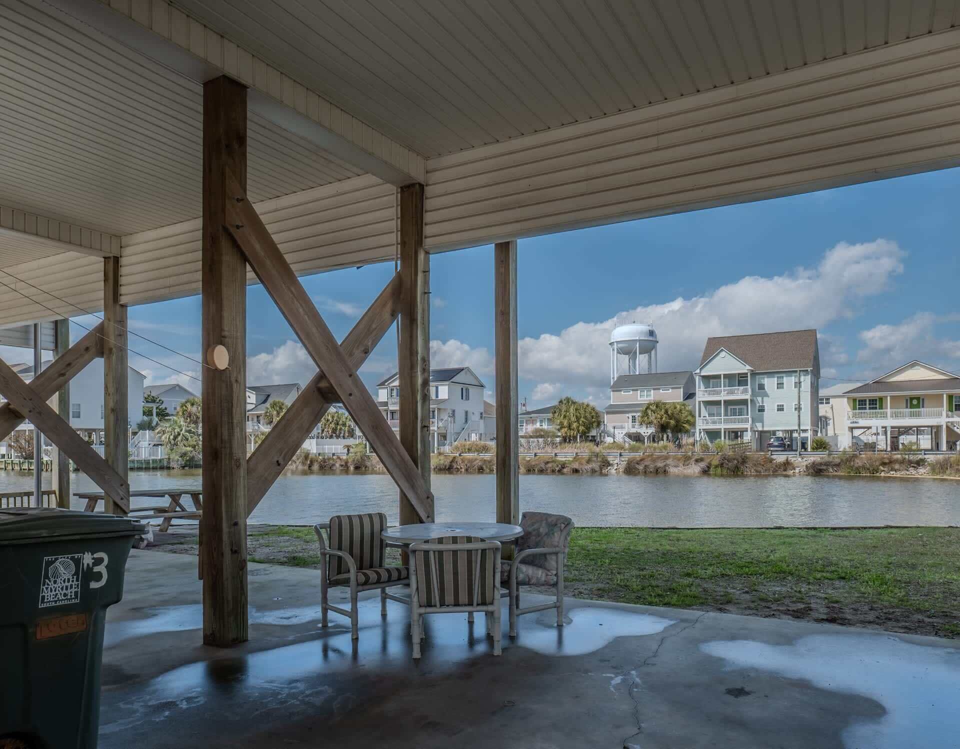 Un espace extérieur abrité, doté d’un sol en béton, de poutres en bois et offrant une vue sur des maisons et un château d’eau situés de l’autre côté de l’eau.