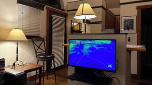 Una habitación con televisión mostrando un pronóstico del tiempo, una mesa con lámpara, una silla y una ventana con persianas.