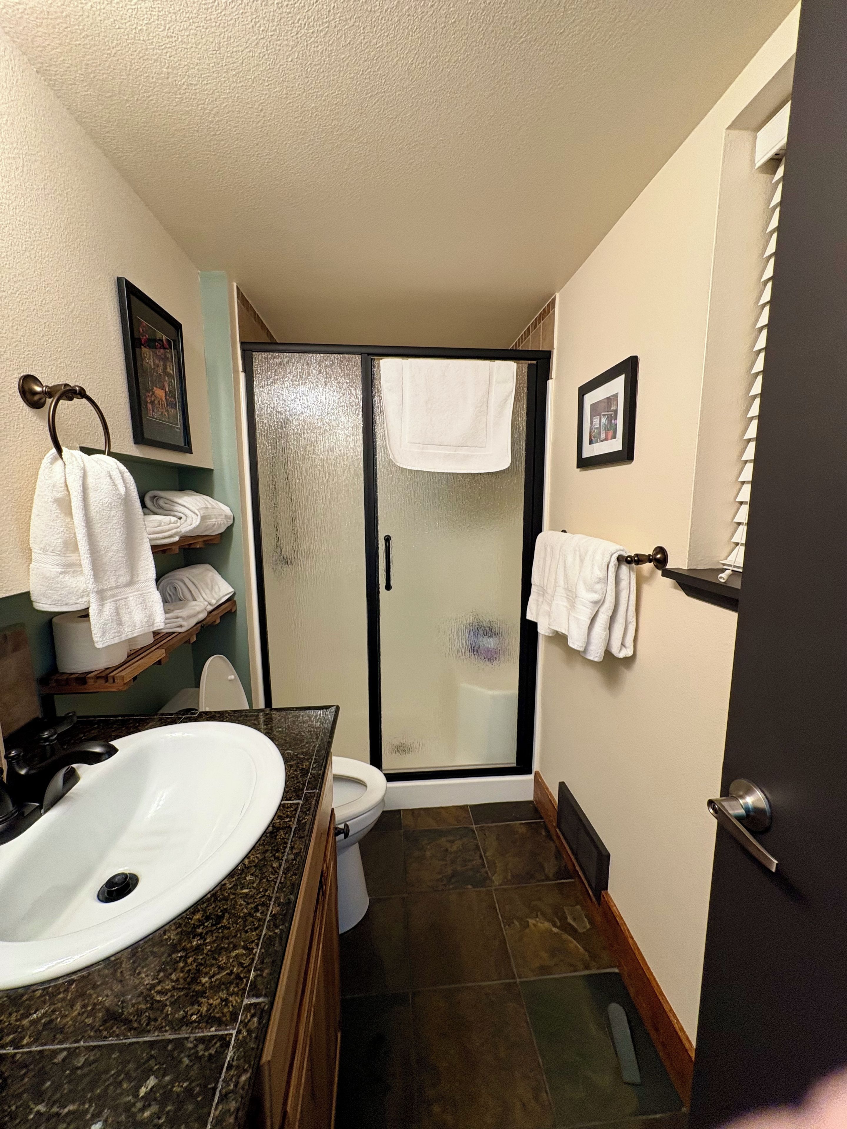 Een badkamer met een wit wastafel, toilet en douchegedeelte. Handdoeken hangen aan een rek en er is een deur met een ruit in.