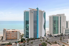 Un hotel moderno con un diseño vertical llamativo, vistas a la playa y una zona de estacionamiento con palmeras.