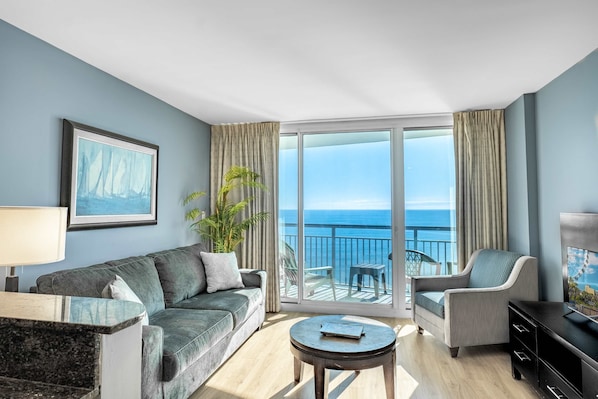 A modern living room with a sofa, armchair, coffee table, and a large window offering a view of the sea.