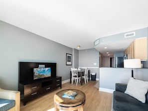 A modern living room with a flat-screen TV, a dining area with chairs, a sofa, and a kitchen in the background.
