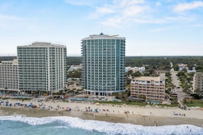 A coastal hotel with multiple floors, a beach in the foreground, and surrounding buildings.