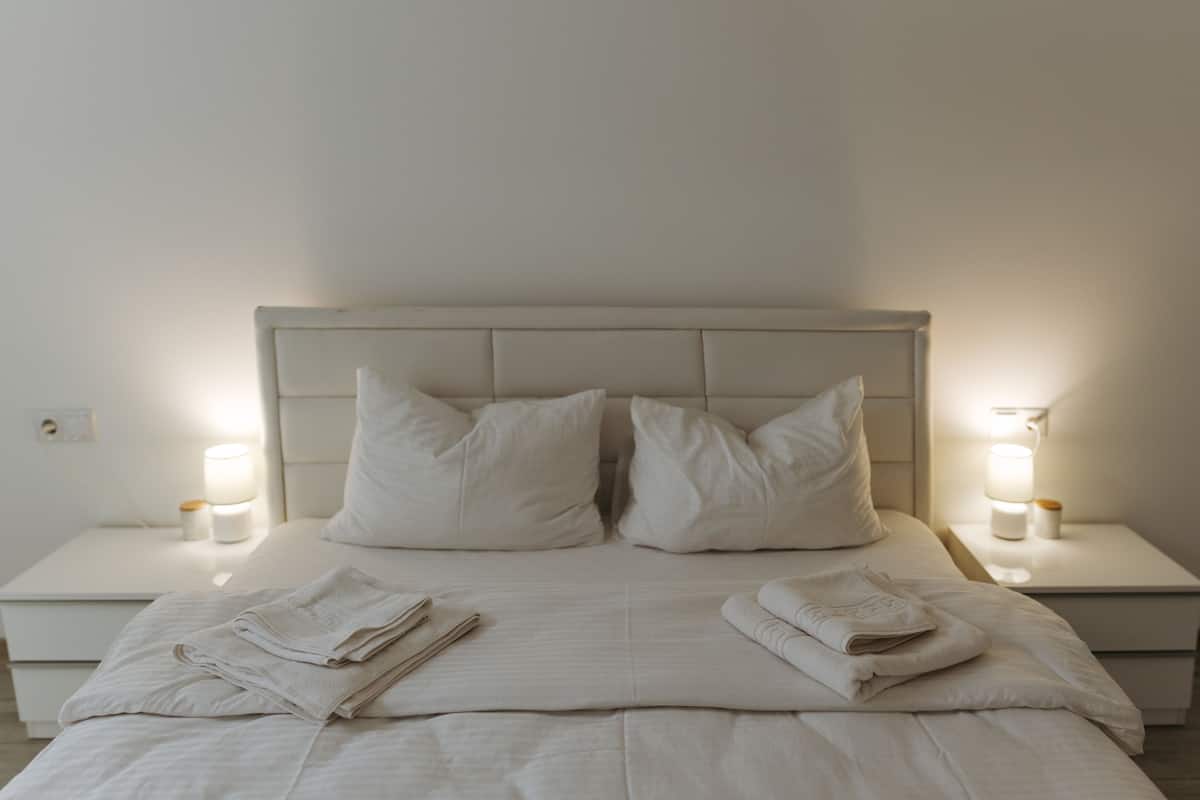 Cama bem arrumada com almofadas e toalhas dobradas, duas luminárias de cabeceira e um criado-mudo branco.