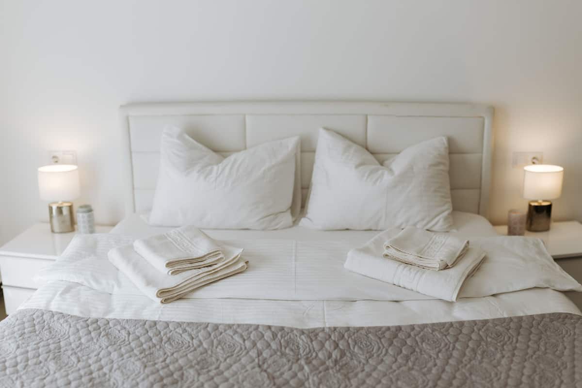 Cama bem arrumada com lençóis brancos, duas luminárias de cabeceira e um painel de cabeceira branco.