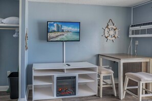 A room with a TV mounted on the wall, a small fireplace, a wooden table, and a chair.
