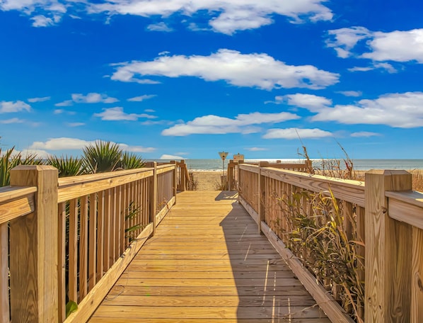 Una pasarela de madera que lleva a una playa, con cielo despejado y un letrero que indica la presencia de socorristas.