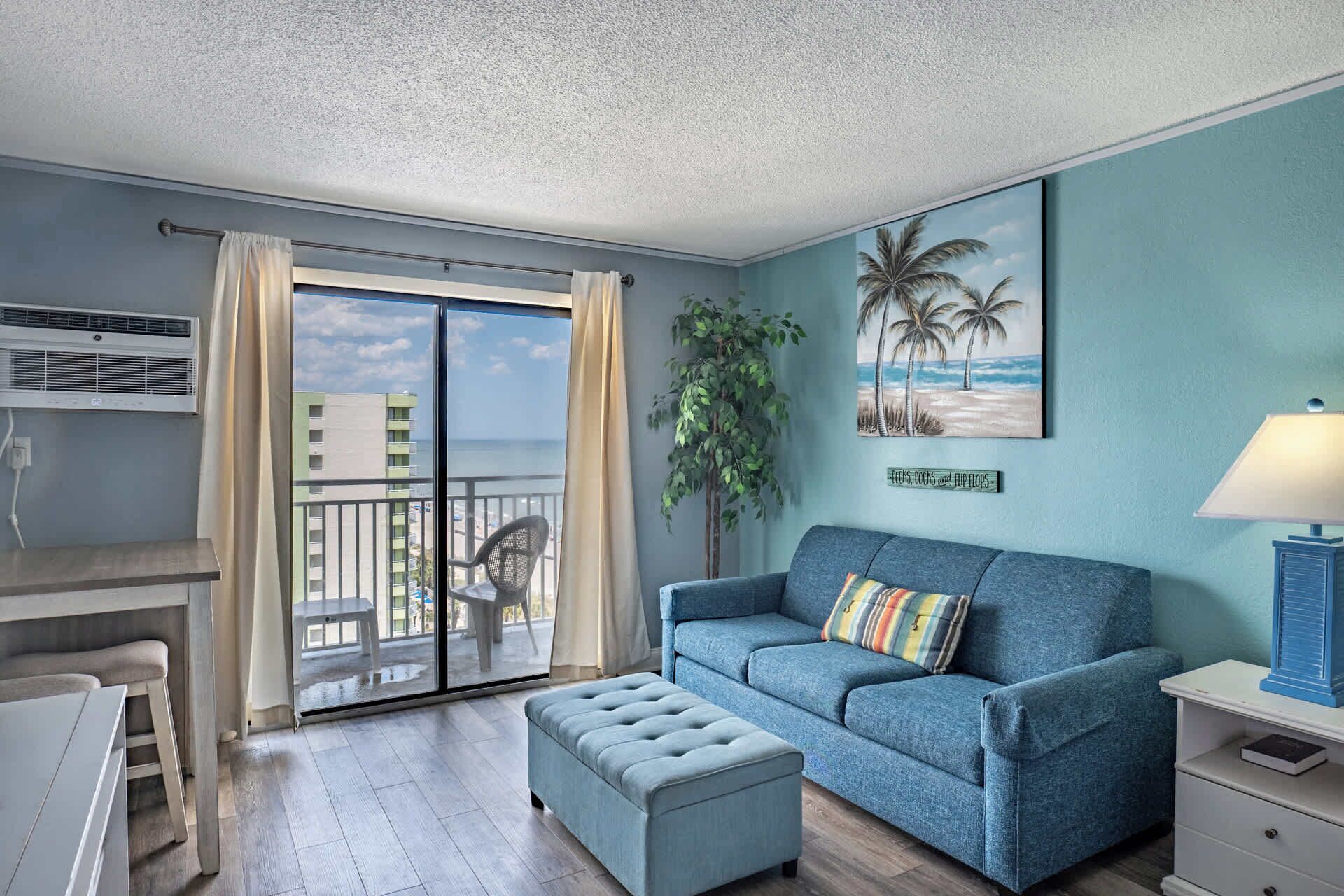 A room with a balcony, a sofa, a chair, a table, and a plant.