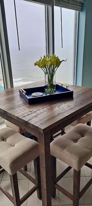 A wooden dining table with a vase of yellow flowers, a tray with plates, and a view of the ocean through large windows.