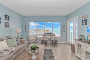 A living room with a sofa, a dining table, and chairs, a potted plant, and a view of the ocean through large windows. - Palace 1810 (Myrtle Beach)