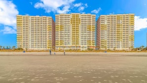 Un hotel moderno frente a la playa con varios balcones, una playa arenosa en primer plano y palmeras.