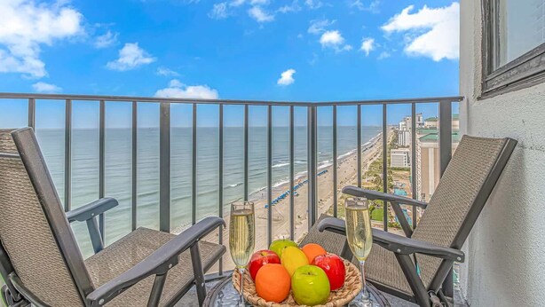 A balcony with wicker chairs, a table with fruit and a glass of champagne, overlooking a beach and ocean. - Palace 1505 (Myrtle Beach)