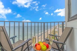 Un balcon avec des fauteuils en osier, une table garnie de fruits et une coupe de champagne, donnant sur une plage et l’océan.