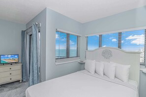 A bedroom with a bed, a TV on a dresser, and a view of the ocean through the windows.