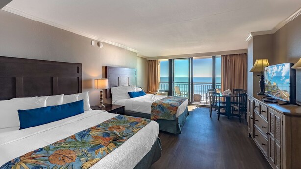 A hotel room with two beds, a TV, a desk with a chair, and a balcony with a view of the ocean. - Caravelle 422 (Myrtle Beach)