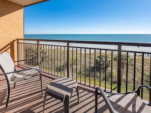 A balcony with a view of the beach, a chair, and a small table.