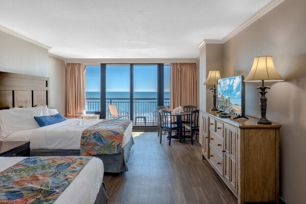 A hotel room with a bed, a desk, a chair, a dresser, and a large window with a view of the ocean.