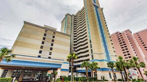 A multi-story hotel with a prominent central tower, a lower building with a blue and beige facade, and palm trees in the foreground. - Carolinian 731 (Myrtle Beach)