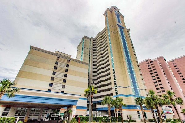 Un hotel de varios pisos con una torre central destacada, un edificio más bajo de fachada azul y beige, y palmeras en primer plano.