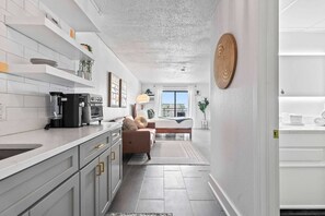 A modern kitchen with white cabinetry, a coffee maker, and a dining area with a sofa and a view of the outdoors.