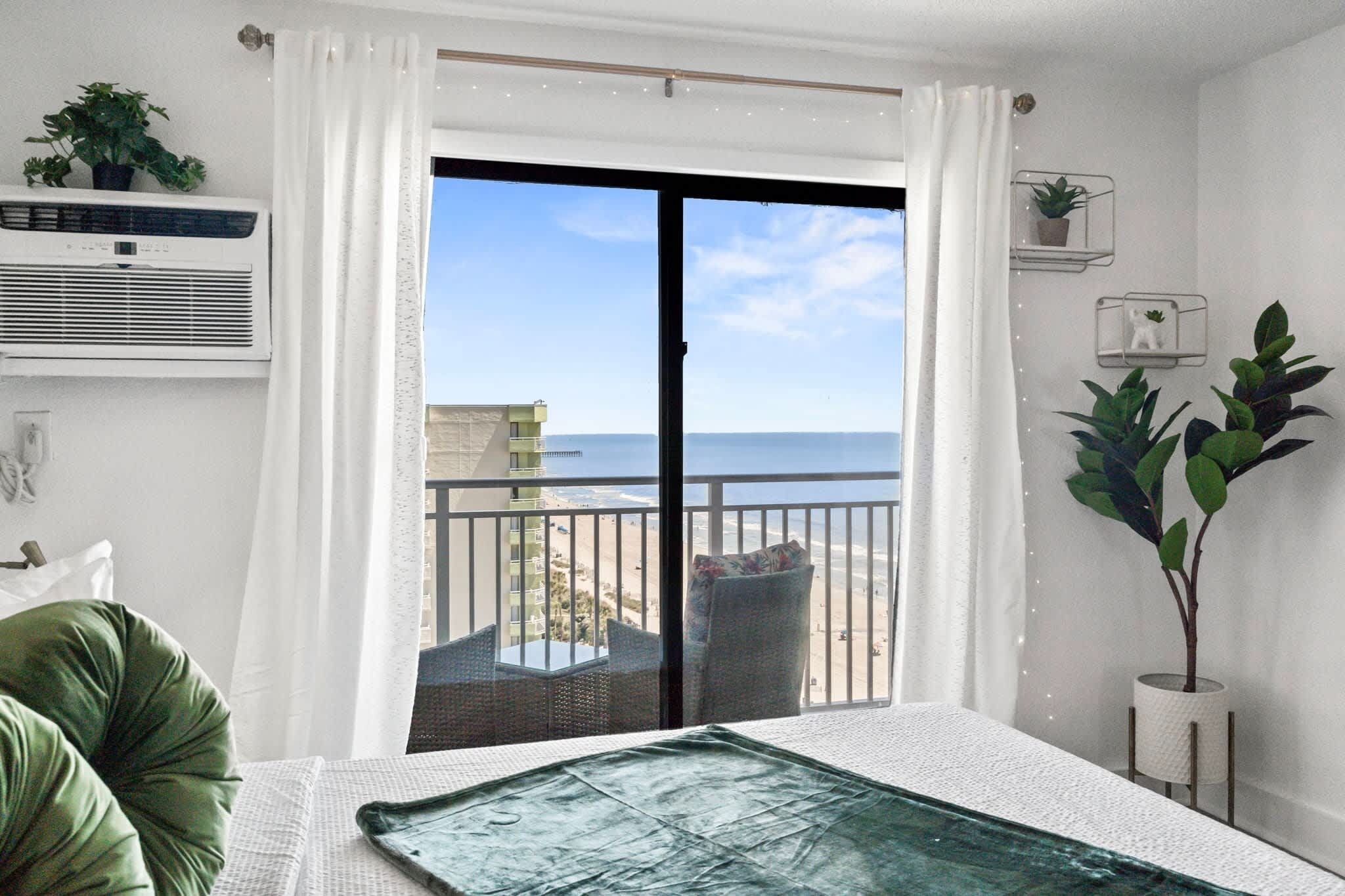 A room with a large sliding glass door leading to a balcony with a view of the beach and ocean.