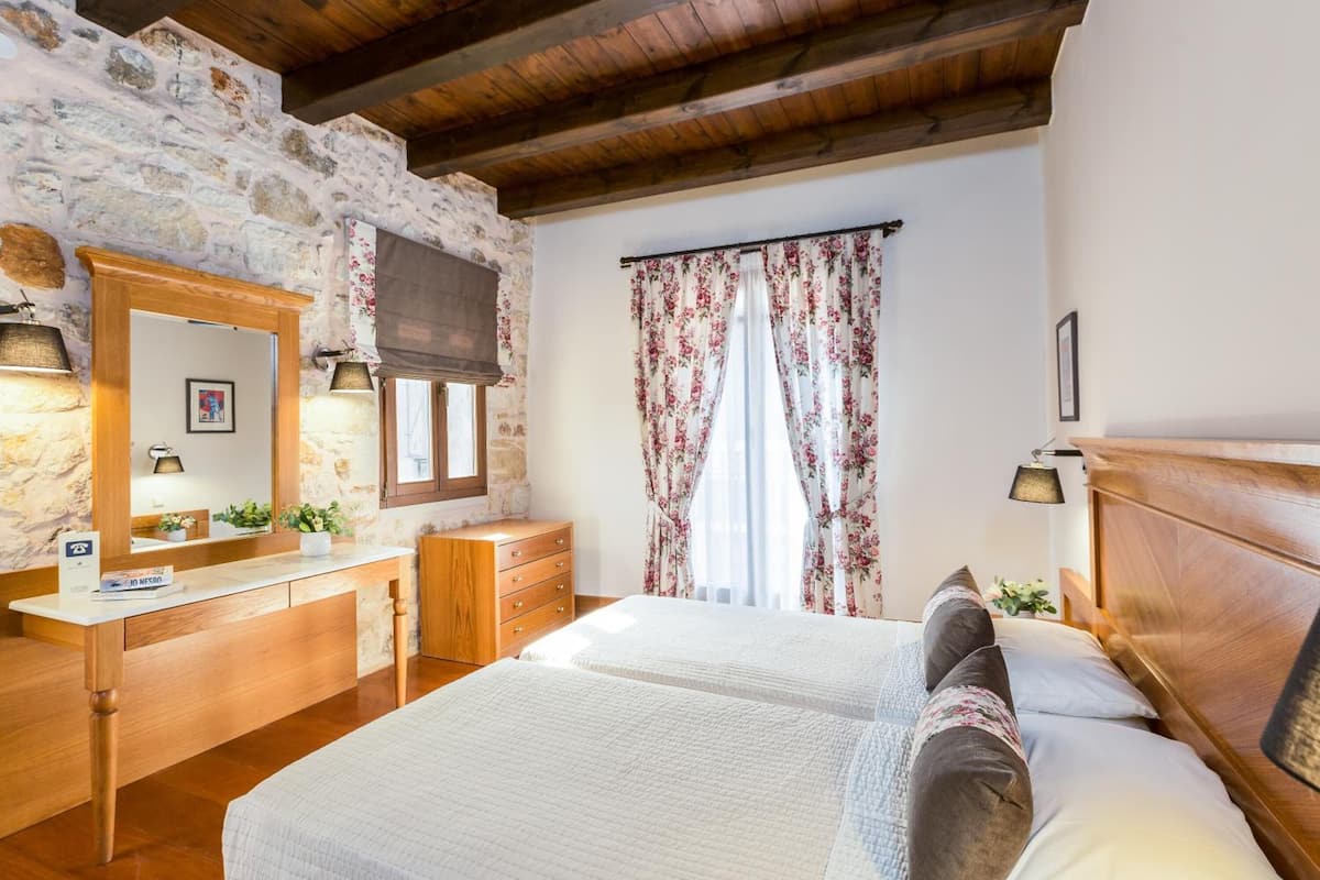Quarto com parede de pedra, vigas de madeira no teto, cama com roupa de cama branca, cómoda de madeira e um espelho.