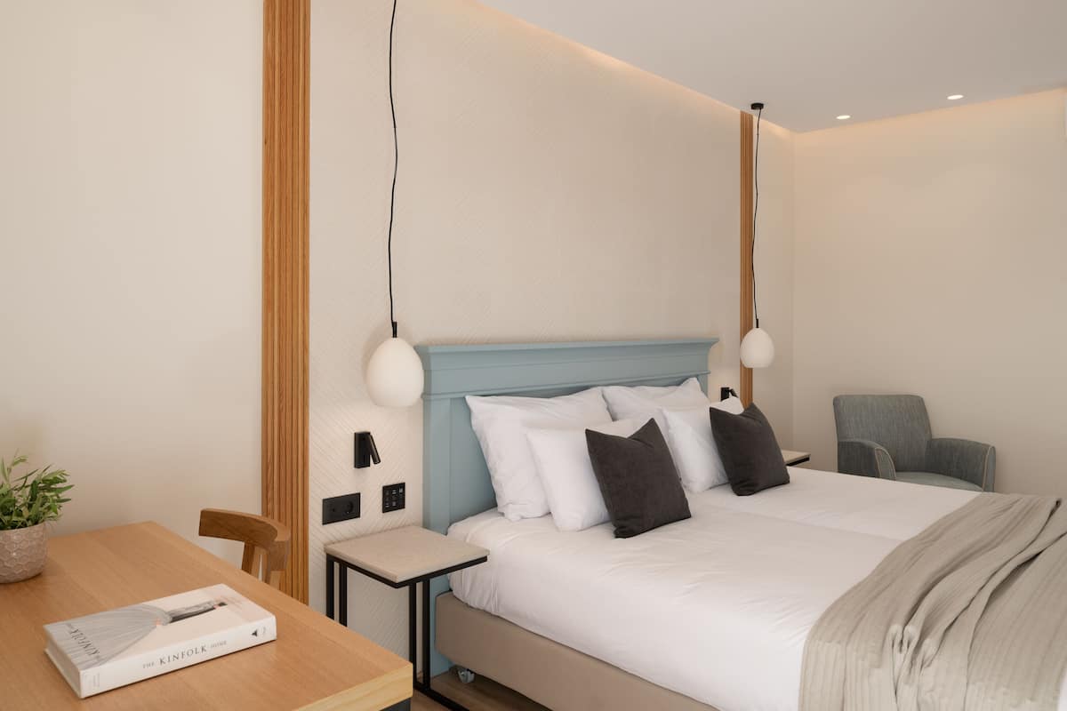 Quarto de hotel moderno com cama, mesa de cabeceira, cadeira e uma pequena mesa com um livro.