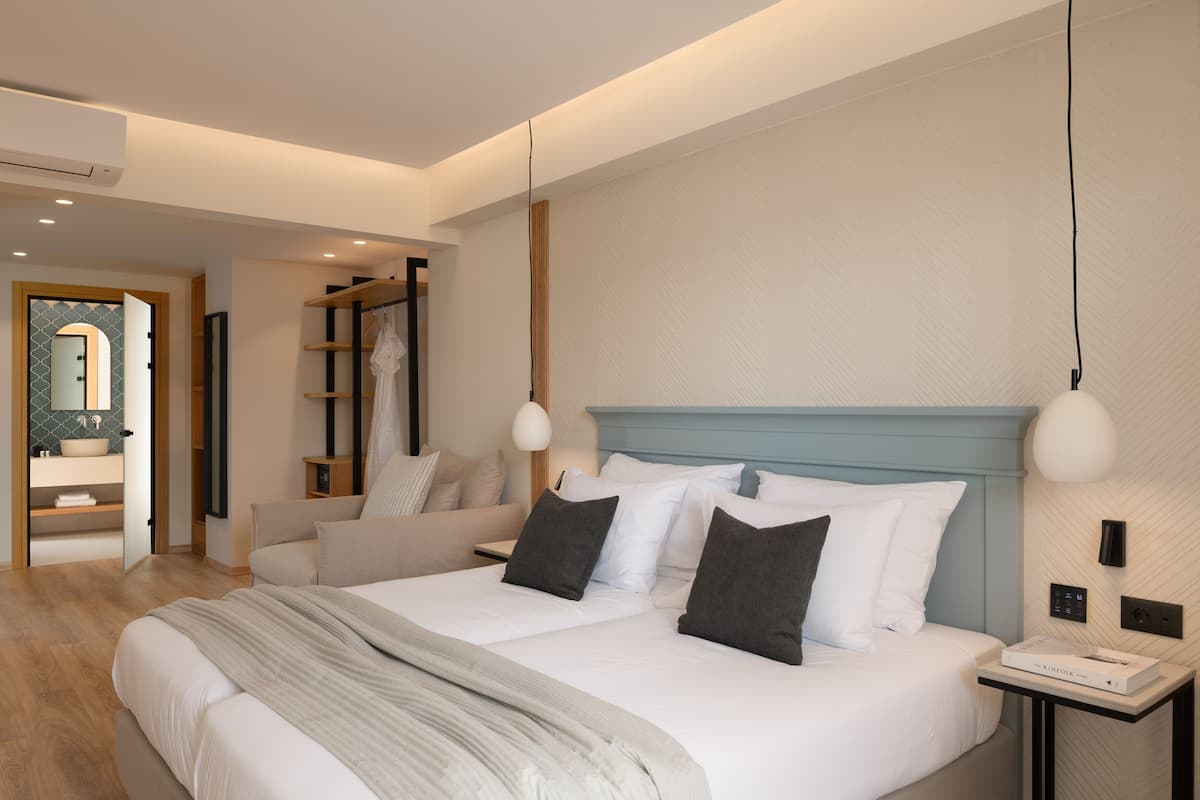 Quarto de hotel moderno com uma cama grande, mesas de cabeceira, um guarda-roupa e um banheiro com espelho e pia.
